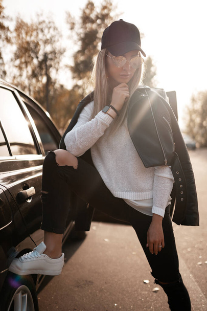 Beauty portret van stijlvolle sexy leuke sport meisje in jas, broek, zonnebril, hoed, sneakers poseren in de buurt van auto op stedelijke achtergrond. Mooie trendy modieuze vrouw model op straat - Foto, afbeelding