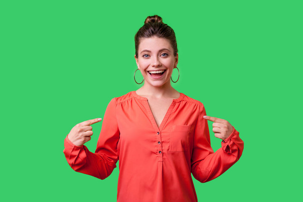 successful woman with bun hairstyle and big earrings in red blouse pointing at herself and looking at camera with proud face on green background - Zdjęcie, obraz