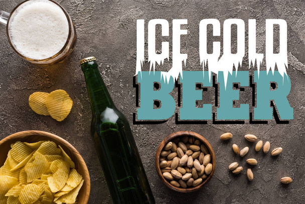 Şişenin yanında şam fıstığı ve cips olan kaselerin üst görüntüsü ve buz gibi bira harfleriyle gri yüzeyde bir kupa bira. - Fotoğraf, Görsel