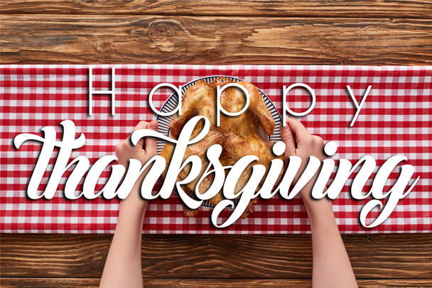 cropped view of woman holding roasted turkey on red plaid napkin on wooden table with happy thanksgiving illustration - Fotoğraf, Görsel