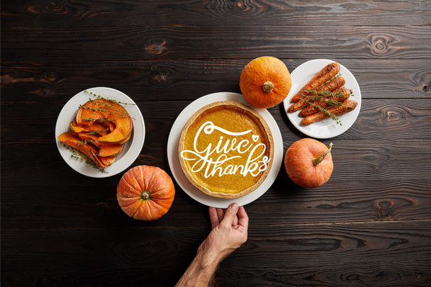 partial view of male hand near plate with pumpkin pie and give thanks illustration, baked whole carrot, sliced and whole pumpkins on dark wooden table - Photo, Image