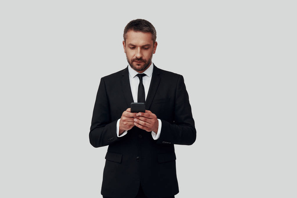 Charming young man in full suit using smart phone while standing against grey background - Zdjęcie, obraz