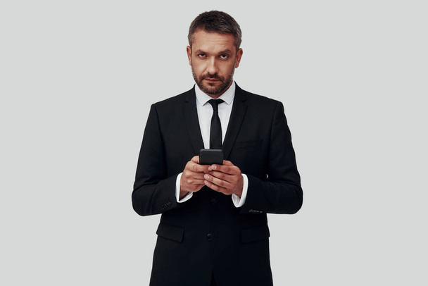 Jovem encantador em terno completo usando telefone inteligente enquanto está de pé contra fundo cinza
 - Foto, Imagem