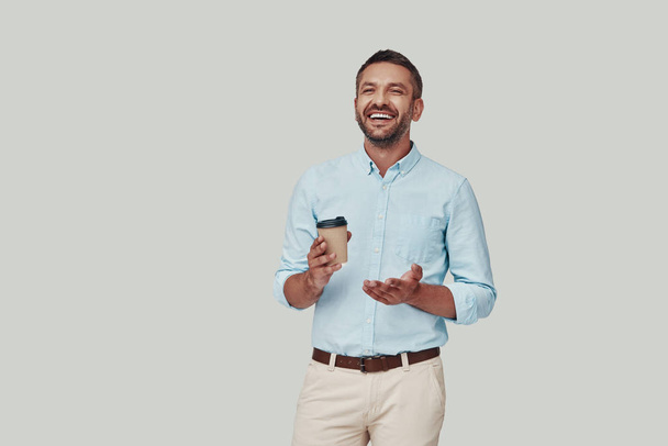 Handsome young man enjoying hot drink and smiling while standing against grey background - Φωτογραφία, εικόνα