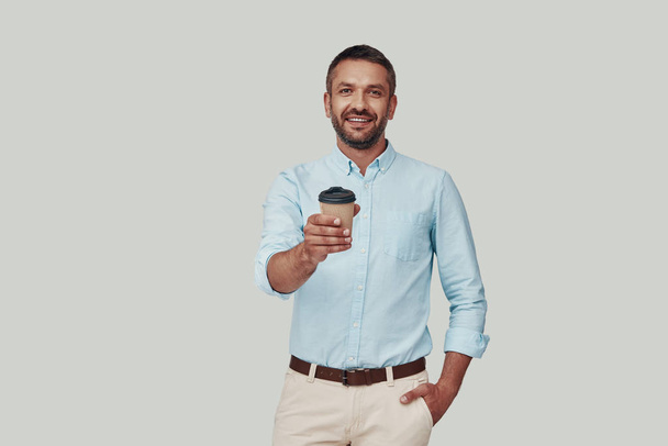 Handsome young man giving you drink and smiling while standing against grey background - Foto, imagen