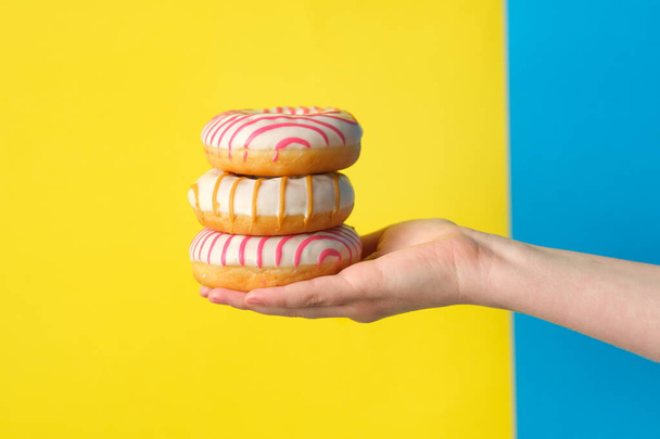 female hand holding pile of sweet donuts on blue and yellow background - Foto, Imagen