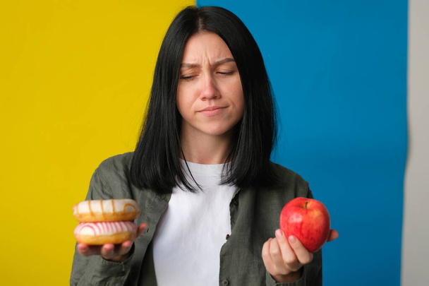 indecisive woman holding donuts and fresh red apple on blue and yellow background - 写真・画像