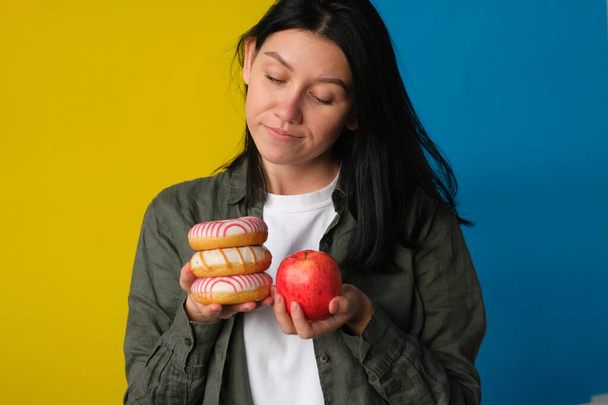 indecisive young woman holding donuts and fresh red apple on blue and yellow background - Фото, изображение