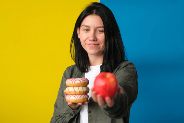 pretty brunette holding donuts and red apple in hands on blue and yellow background - Zdjęcie, obraz