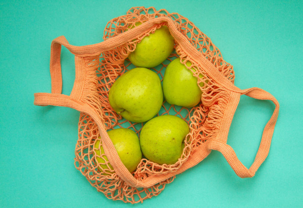 As maçãs estão em um saco de corda laranja claro. Malha saco de compras com frutas em um fundo de menta neo
.  - Foto, Imagem