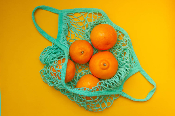 Tangerines in green mint color string bag on a turquoise background. Citrus in a wicker bag on paper. Shot from above. - Photo, Image