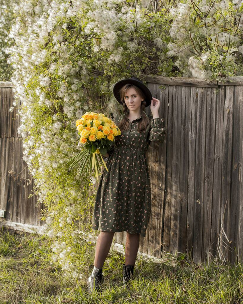 Beautiful girl with a bouquet of yellow roses. Woman with model appearance in a hat. Pretty woman near a vintage fence. Teenager girl in a black hat and roses. - 写真・画像