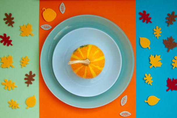 Decorative beautiful bright pumpkins on a paper background of their three colors - orange, blue and green. Paper cut autumn leaves around a plate with pumpkin. - Фото, изображение