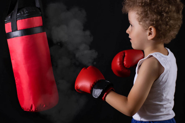 Ein kleiner Junge in roten Boxhandschuhen trainiert, den Boxsack zu treffen. Sport, Kinderboxen. - Foto, Bild