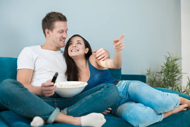 young couple beautiful brunette woman and handsome man watching movie on the sofa eating pop corn she shows him exciting moment by pointing her finger - Photo, image