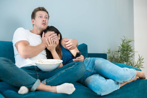 young couple beautiful brunette woman and handsome man watching scary movie on the sofa eating pop corn looking frightened he closes her eyes homesters - Φωτογραφία, εικόνα