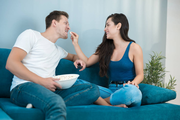 young couple beautiful brunette woman and handsome man watching movie on the sofa wife feeds husband with pop corn homesters - Foto, Imagen