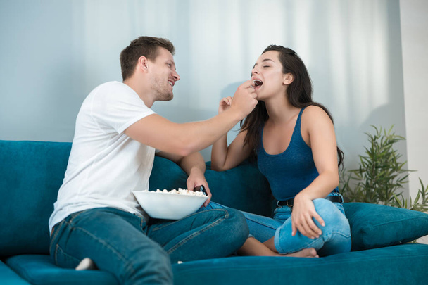 Nuori pari kaunis brunette nainen ja komea mies katsomassa elokuvaa sohvalla aviomies ruokkii vaimonsa pop maissi kodeissa
 - Valokuva, kuva