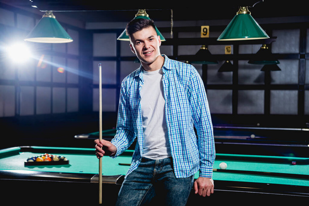 Young man playing in billiard. Posing near the table with a cue in his hands. Pool background. - Foto, afbeelding