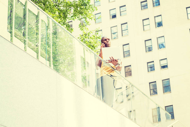 Trabajando en cualquier lugar. Joven afroamericano que trabaja en la ciudad de Nueva York, parado junto a una cerca de cristal frente a un edificio de oficinas en el distrito de negocios, hablando por teléfono celular, trabajando en una computadora portátil
 - Foto, Imagen