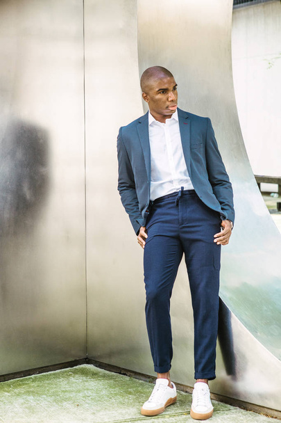 Portret van de jonge Afro-Amerikaanse man in New York City. Het dragen van blauwe blazer, wit shirt, blauwe broek, witte sneakers, jonge zwarte student staande tegen zilveren metalen muur op de campus, denk na - Foto, afbeelding
