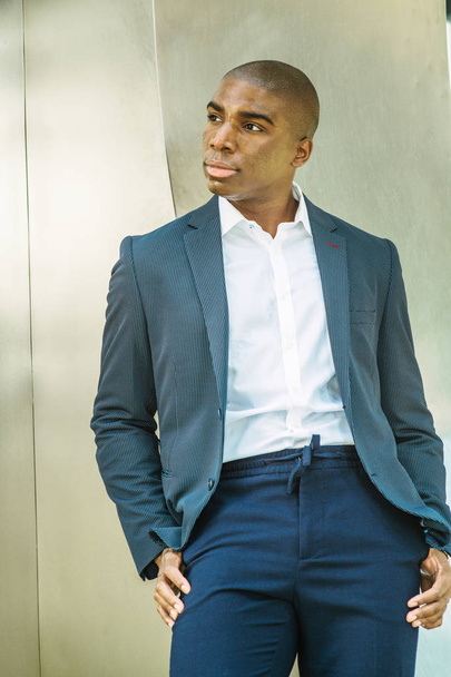 Portrait of Young African American Man in New York City. Wearing blue blazer, white shirt, blue pants, young black college student standing against silver metal wall on campus, looking away, thinking. - Photo, Image