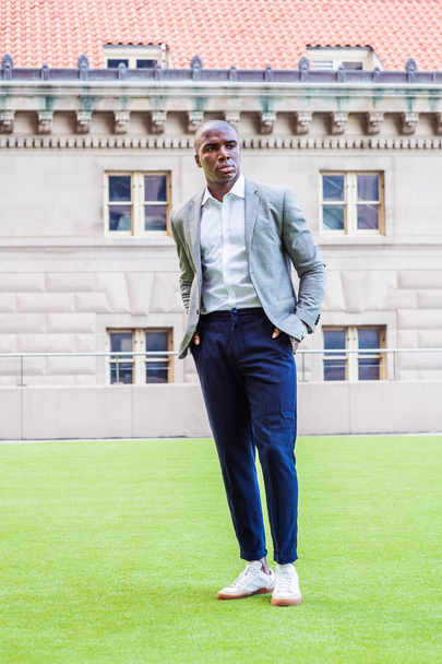 Young African American Man Street Fashion in New York City. Black college student with short hair, wearing gray patterned blazer, white shirt, blue pants, sneakers, standing on green lawn on campus - 写真・画像