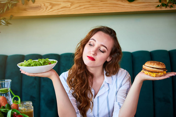 Beautiful young woman decides eating hamburger or fresh salad in kitchen. Cheap junk food vs healthy diet - Фото, изображение
