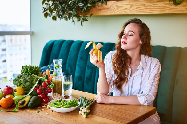 美しい若い女性食べるバナナ座ってキッチン - 写真・画像