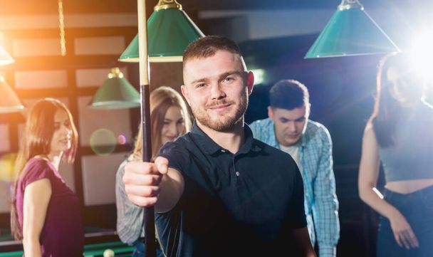 Young man playing in billiard. Posing near the table with a cue in his hands. His friends on the background. - Foto, afbeelding