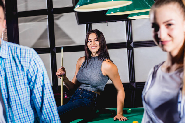 Young woman playing in billiard. Posing near the table with a cue in her hands. A group of friends on the background. - Photo, Image