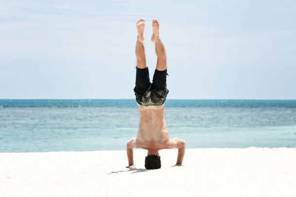 Young man in excellent physical shape headstands on the beach - Foto, immagini