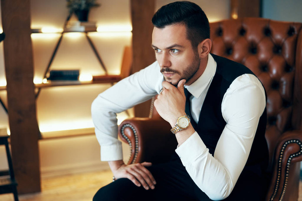 Handsome young man sitting and posing in armchair in modern luxury interior. Fashion portrait - 写真・画像