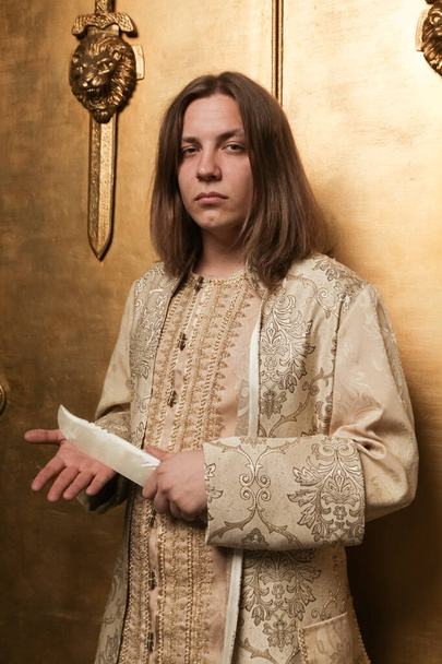 uroczy nieśmiały młody chłopak w średniowiecznym stylu złoty garnitur w studio fotograficznym jest gotowy, aby przejść do partii. Człowiek z nożem zrobionym z nefrytu - Zdjęcie, obraz
