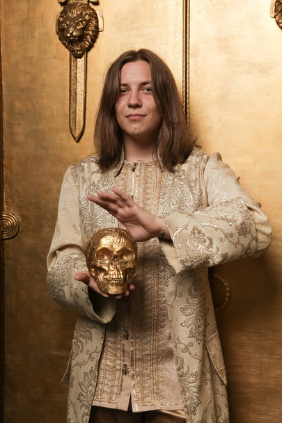 charmante verlegen jonge tiener man in een middeleeuwse stijl gouden pak in een foto Studio is klaar om naar een feest te gaan. man met een grote vergulde menselijke schedel - Foto, afbeelding