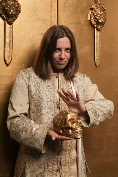 γοητευτικός ντροπαλός έφηβος με μεσαιωνικό χρυσό κοστούμι σε ένα φωτογραφικό στούντιο είναι έτοιμος να πάει σε ένα πάρτι. άνθρωπος με μεγάλο επίχρυσο ανθρώπινο κρανίο - Φωτογραφία, εικόνα