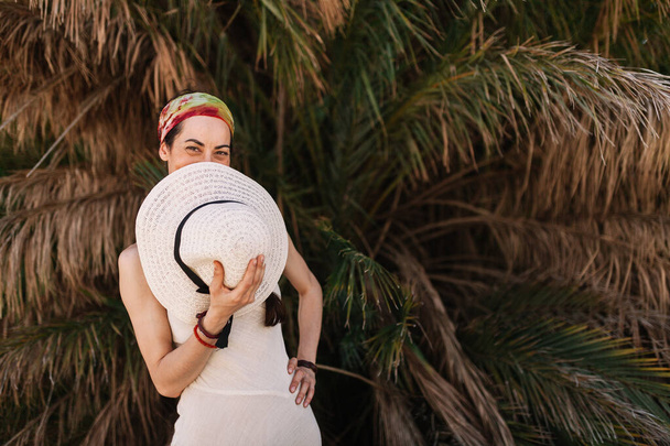 Woman wearing dress standing close to palm tree covers her mouth - Photo, Image