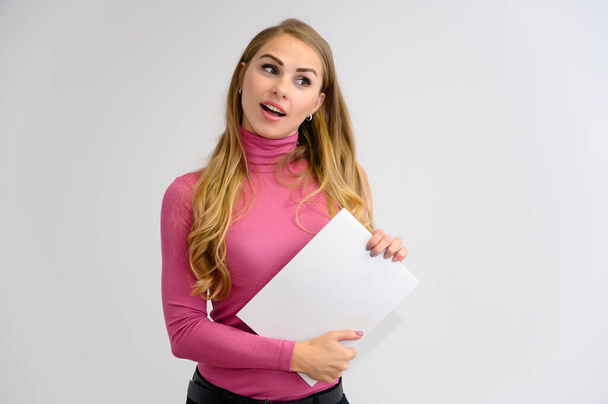 Close-up portrait of a pretty blonde girl with long curly hair standing in the studio on a white background with emotions in different poses in a pink sweater with a folder in her hands. - Фото, зображення