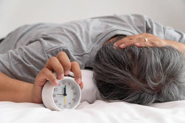 Asiático de mediana edad hombre está sosteniendo un despertador blanco y su cara mostró aburrimiento y sentirse mal, su problema es un trastorno del sueño. El concepto de sufrimiento insomnio
 - Foto, Imagen