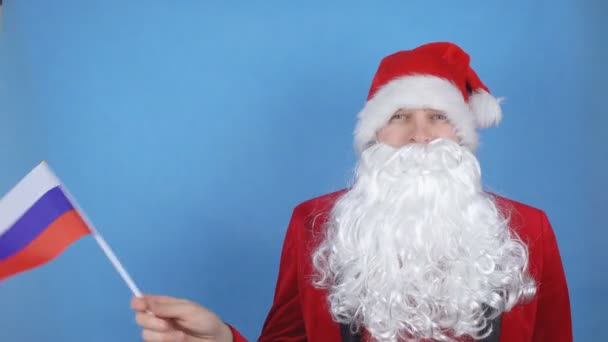 A man in a Santa Claus costume with a beard waving the flag of Russia on a blue background. New Year holidays concept in the world - Video, Çekim