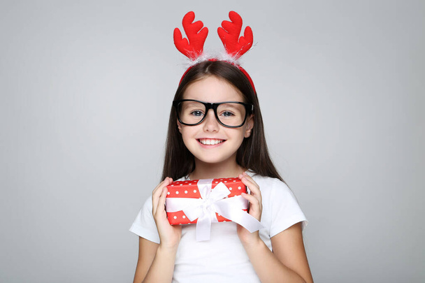 Little girl in christmas red horns holding gift box on grey back - Фото, зображення