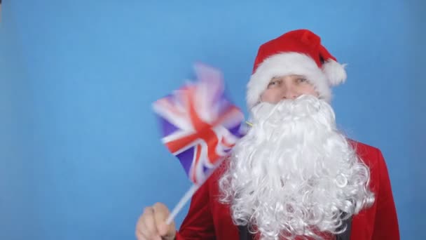 A man in a Santa Claus costume with a beard waving the flag of Britannia on a blue background. New Year holidays concept in the world - Video, Çekim