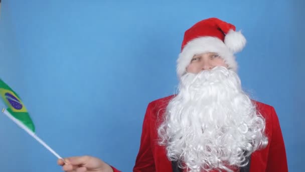 A man in a Santa Claus costume with a beard waving the flag of Brazil on a blue background. New Year holidays concept in the world - Imágenes, Vídeo