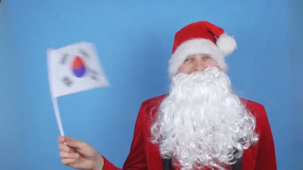 A man in a Santa Claus costume with a beard waving the flag of South Korea on a blue background. New Year holidays concept in the world - Záběry, video