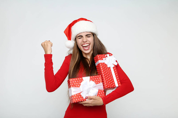Beautiful woman wearing santa hat and holding gift boxes on grey - Photo, image