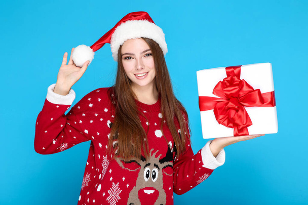 Beautiful woman wearing christmas sweater, santa hat and holding gift box on blue background - Photo, image