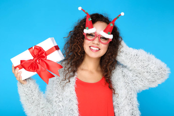 Beautiful american woman in fashion clothing and eyeglasses with gift box on blue background - Zdjęcie, obraz
