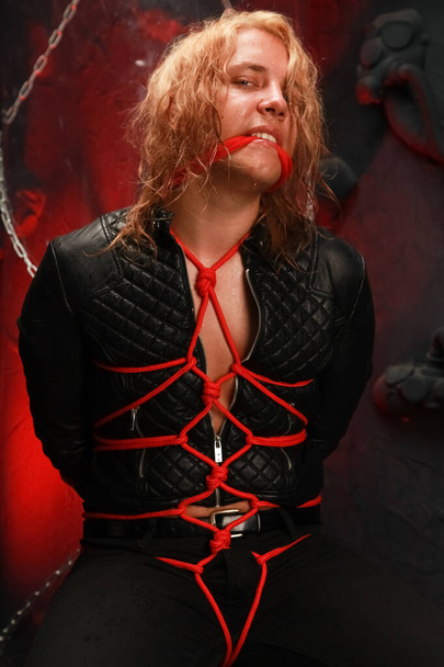handsome young long-haired guy sits in a leather jacket and is tied with red ropes. sexy bondage slave lobes bdsm and be dominated - Foto, imagen