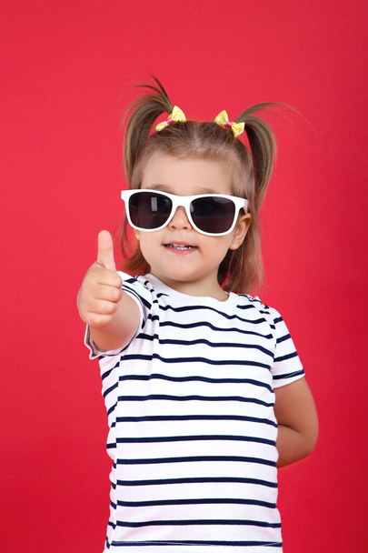 Cute little girl in sunglasses showing thumb up on red backgroun - Foto, Bild