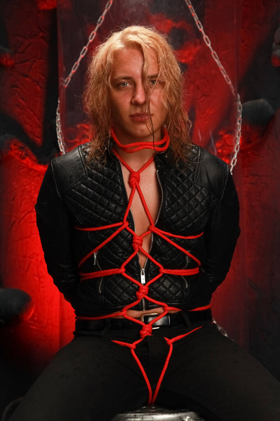 handsome young long-haired guy sits in a leather jacket and is tied with red ropes. sexy bondage slave lobes bdsm and be dominated - 写真・画像